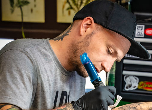 Image centers on a Travis the tattoo artist, captured from the chest up, deeply engrossed with intense concentration in his work.